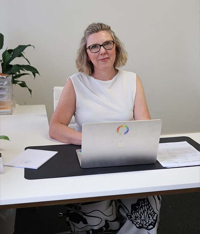 Kylie Anderson Adelaide Nutritionist at her desk on Halifax street. Sitting with a laptop open, smiling with a white top on.